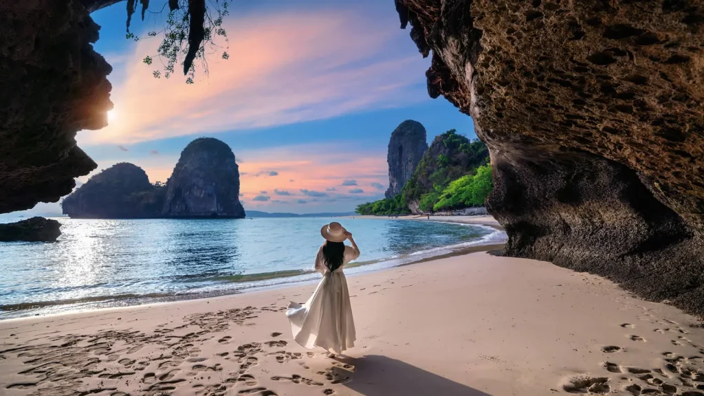 A woman in a white dress stands gracefully on a sandy beach, illuminated by the warm hues of a sunset.