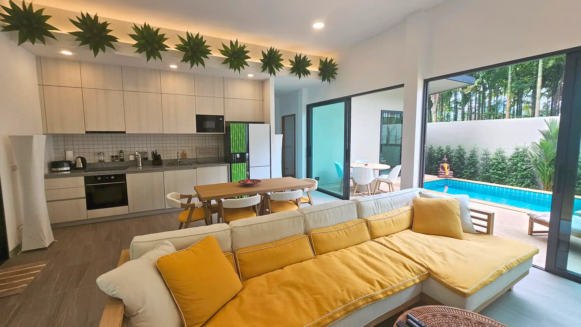 Modern villa bright open-plan living space with modern kitchen, wooden dining table, and lounge area featuring a sectional sofa, opening through sliding glass doors to a patio and swimming pool surrounded by tropical greenery.
