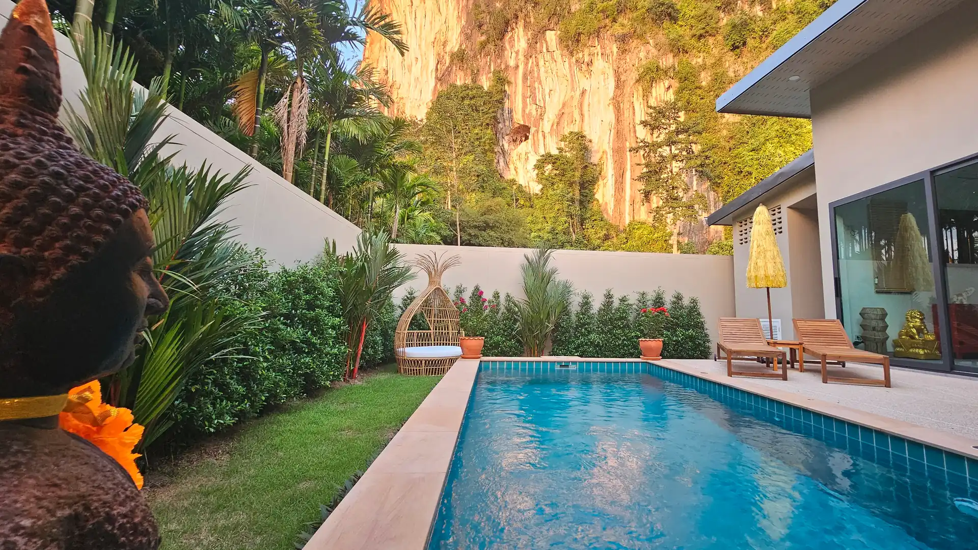 Modern villa private tropical poolside retreat with Buddha statue, yellow umbrella, and lounge chairs, framed by lush greenery and dramatic limestone karst cliff backdrop.