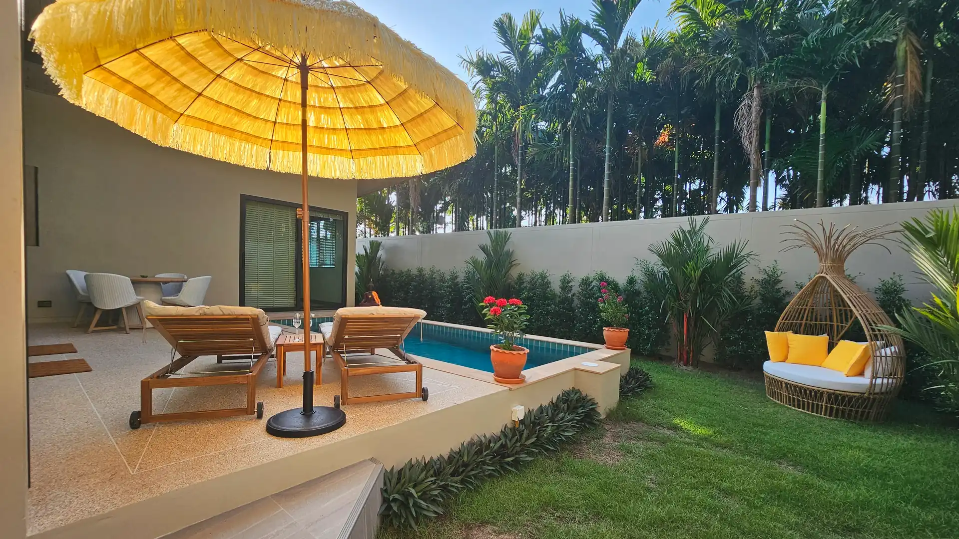 Contemporary villa tropical patio with swimming pool, large yellow patio umbrella, wooden reclining sun loungers, outdoor table and 4 chair set, rattan egg-shaped sofa, and lush palm lined garden.