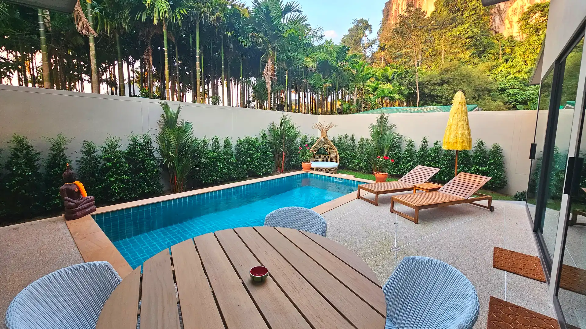Modern villa tropical outdoor area with pool, yellow patio umbrella, reclining wooden sun chairs, round table with blue chairs, and a Buddha statue by lush greenery against limestone cliff backdrop with palms and large native trees.