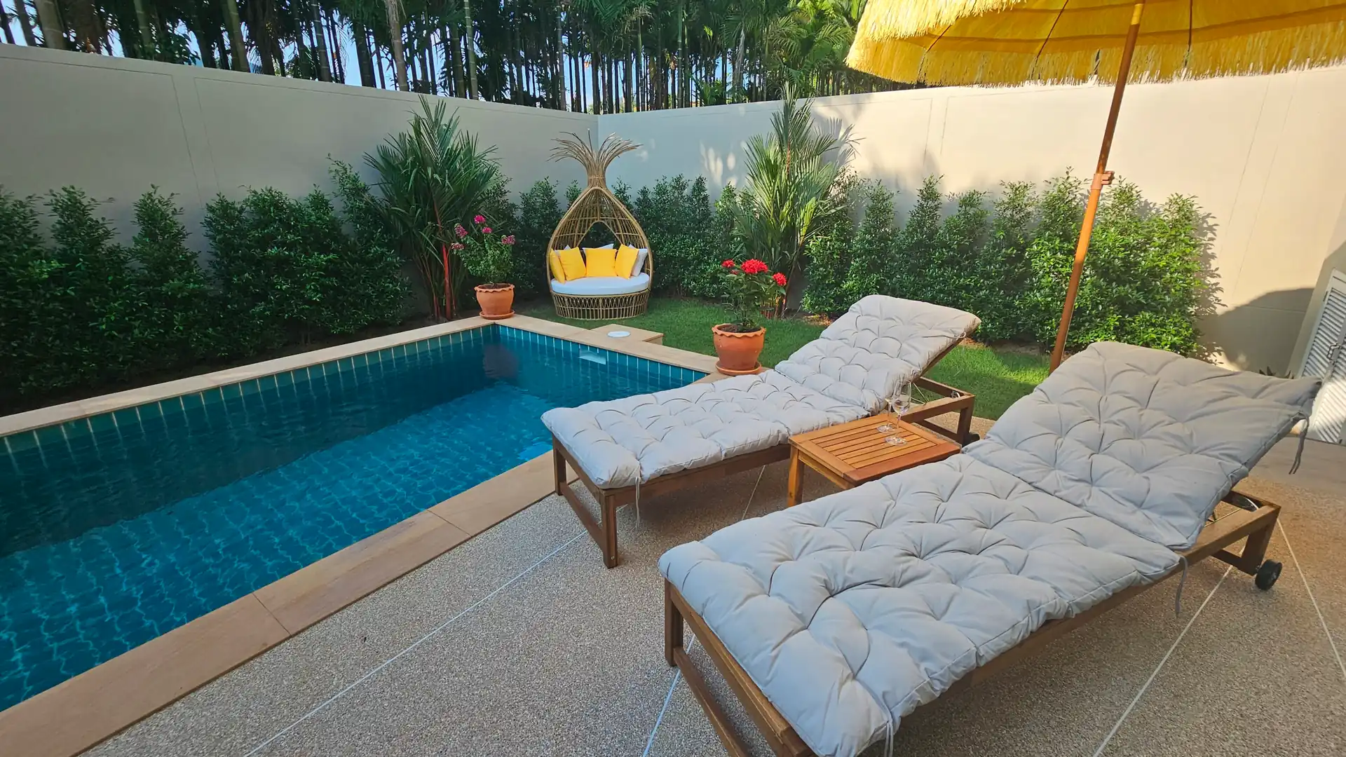Private tropical pool area with cushioned reclining sun loungers under a large yellow umbrella, wicker chair with bright cushions, and lush greenery creating a serene retreat.